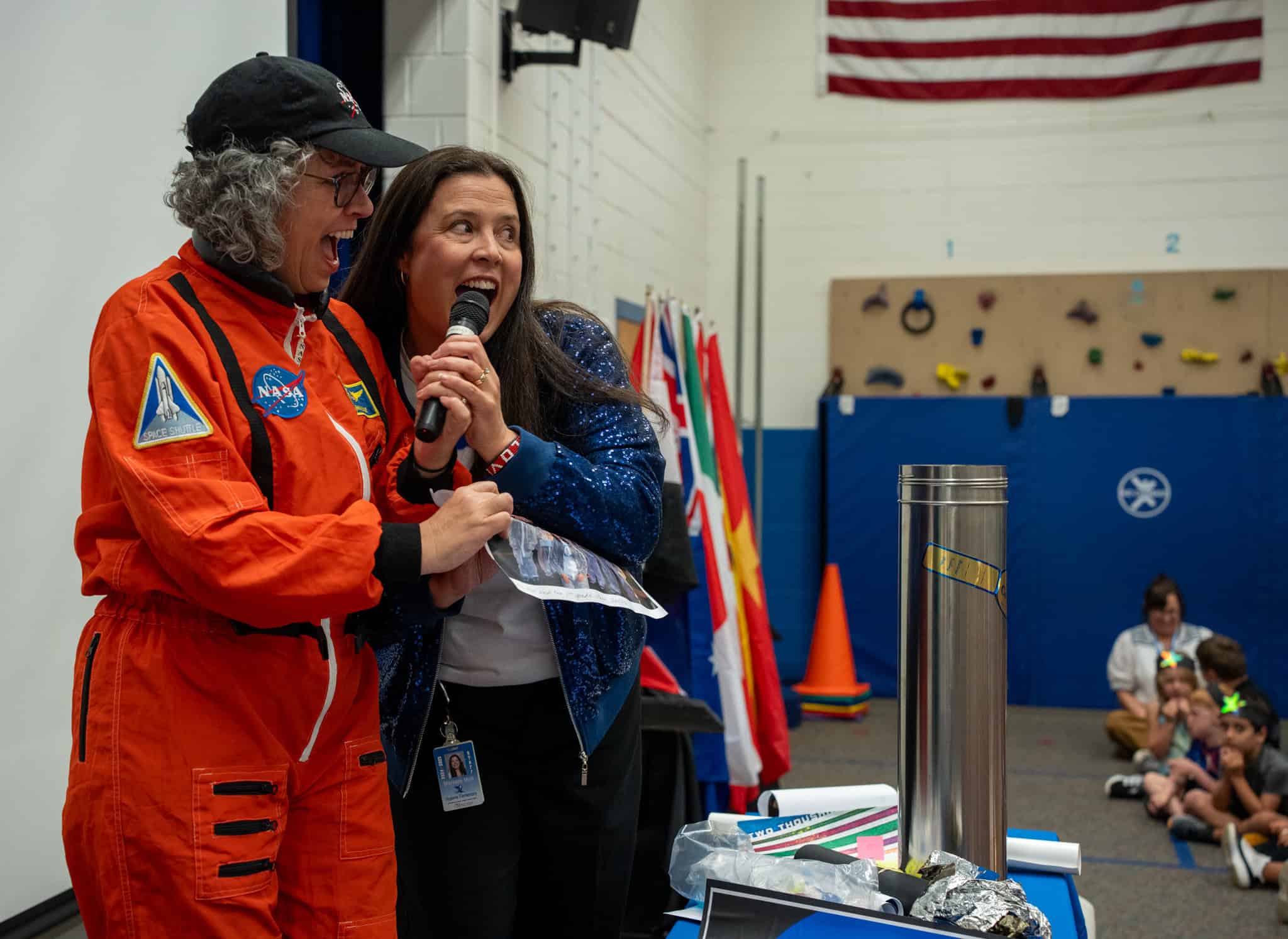 Two women, one in an astronaut suit and the other with a microphone, engage an audience in a school setting.