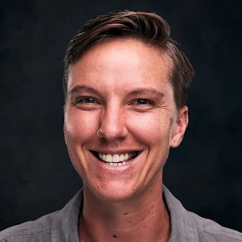 Smiling person with short hair and a nose ring, wearing a gray shirt against a dark background.