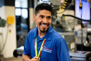 A smiling man in a blue shirt stands in a workshop, surrounded by machinery, wearing a lanyard with ID tags.