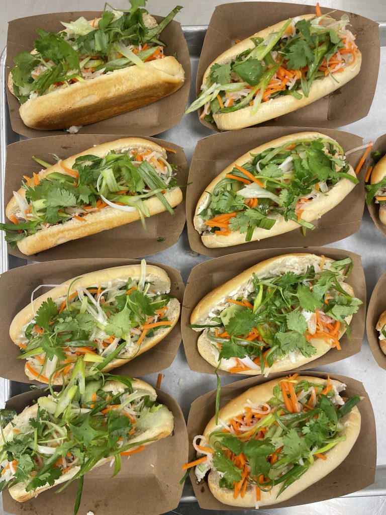 Several sandwiches are arranged in brown paper trays, filled with vegetables and topped with fresh cilantro.