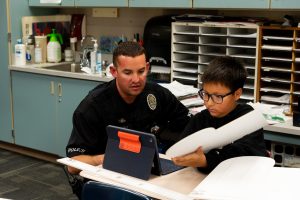 A police officer and a young student collaborate over a tablet in a classroom setting.
