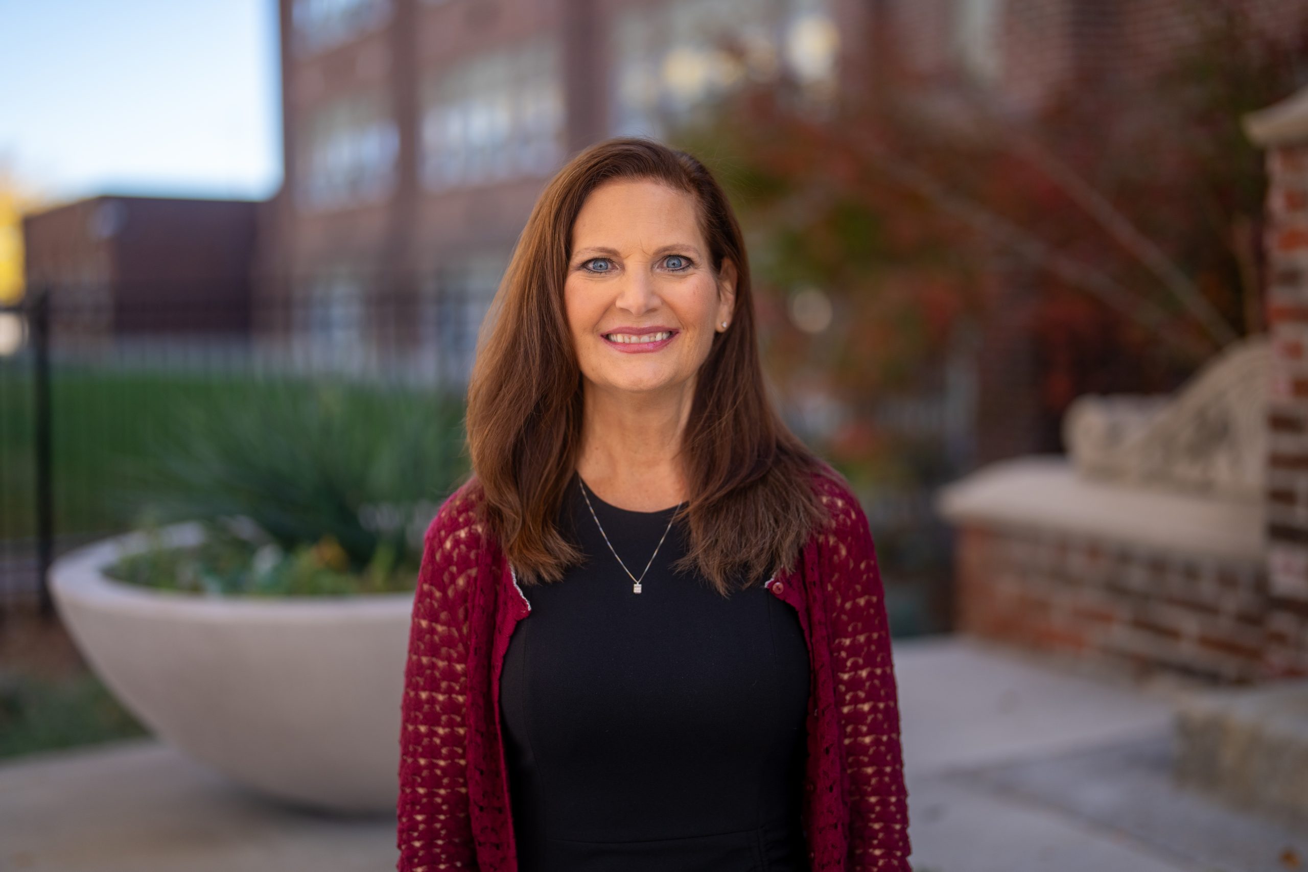 A woman with long brown hair smiles in front of a blurred brick building and greenery, wearing a black dress and maroon cardigan.
