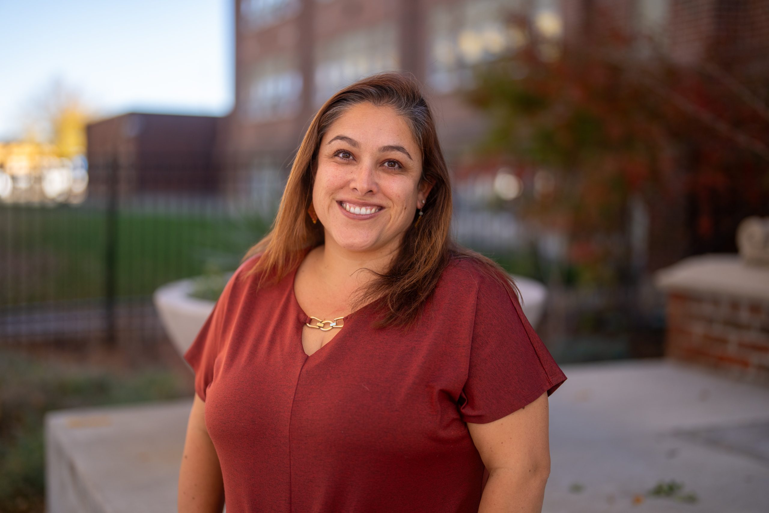 A smiling woman with long hair stands outdoors, wearing a red shirt, with a blurred school building in the background.