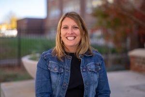 Smiling woman with long hair wearing a denim jacket, sitting outdoors with a blurred background of greenery and buildings.