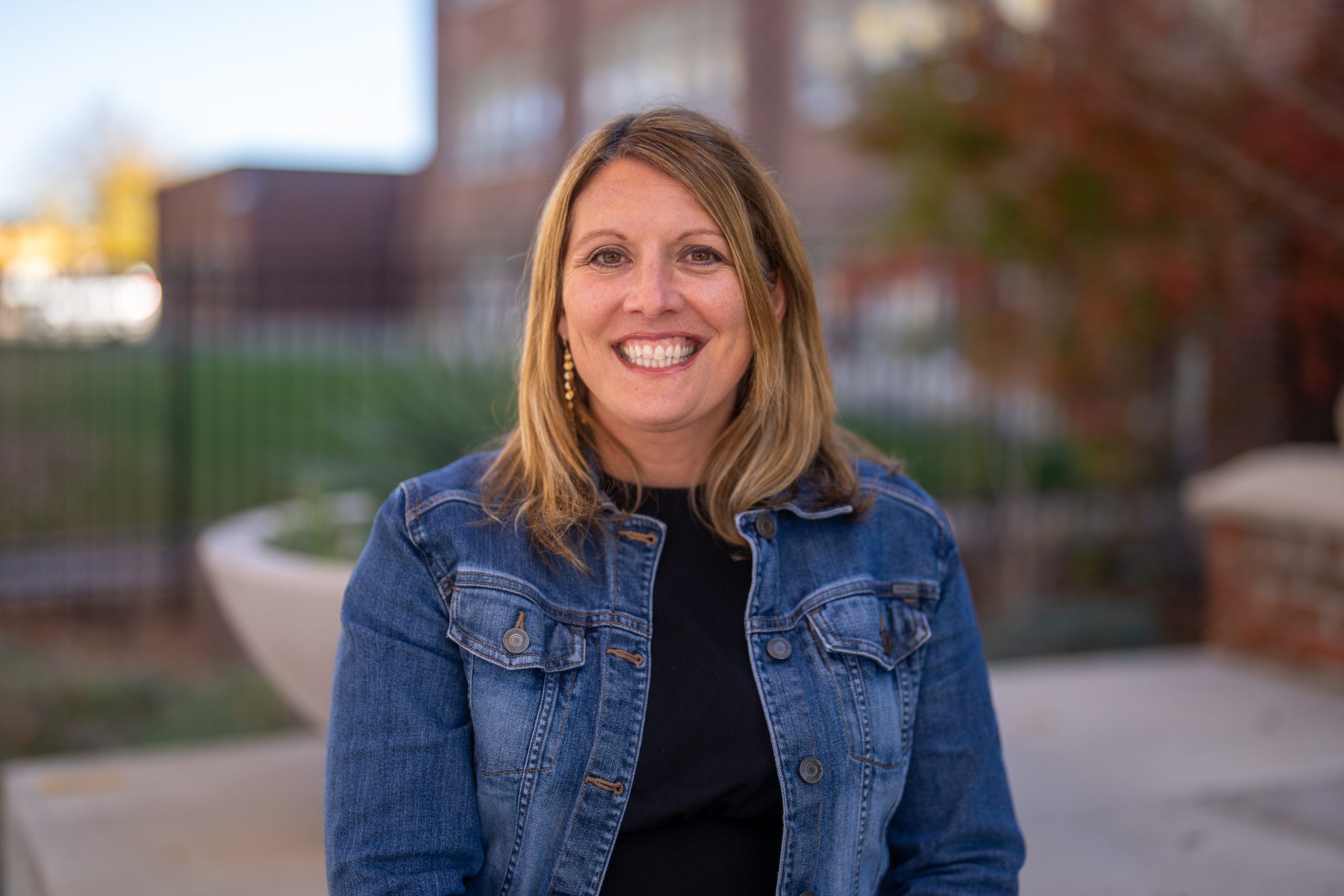 Smiling woman with long hair wearing a denim jacket, sitting outdoors with a blurred background of greenery and buildings.
