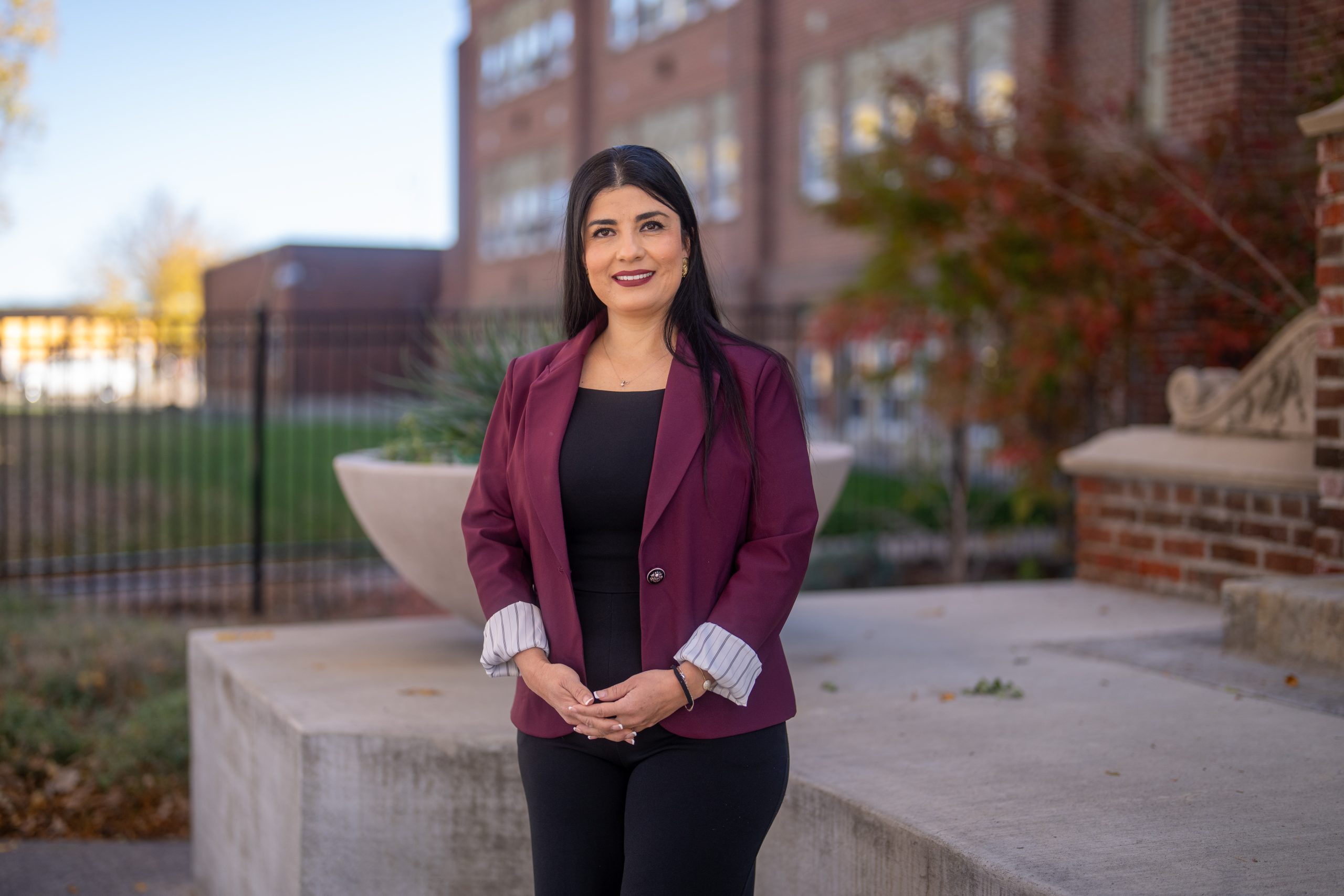 A woman in a maroon blazer stands outdoors, smiling, with a school building and greenery in the background.