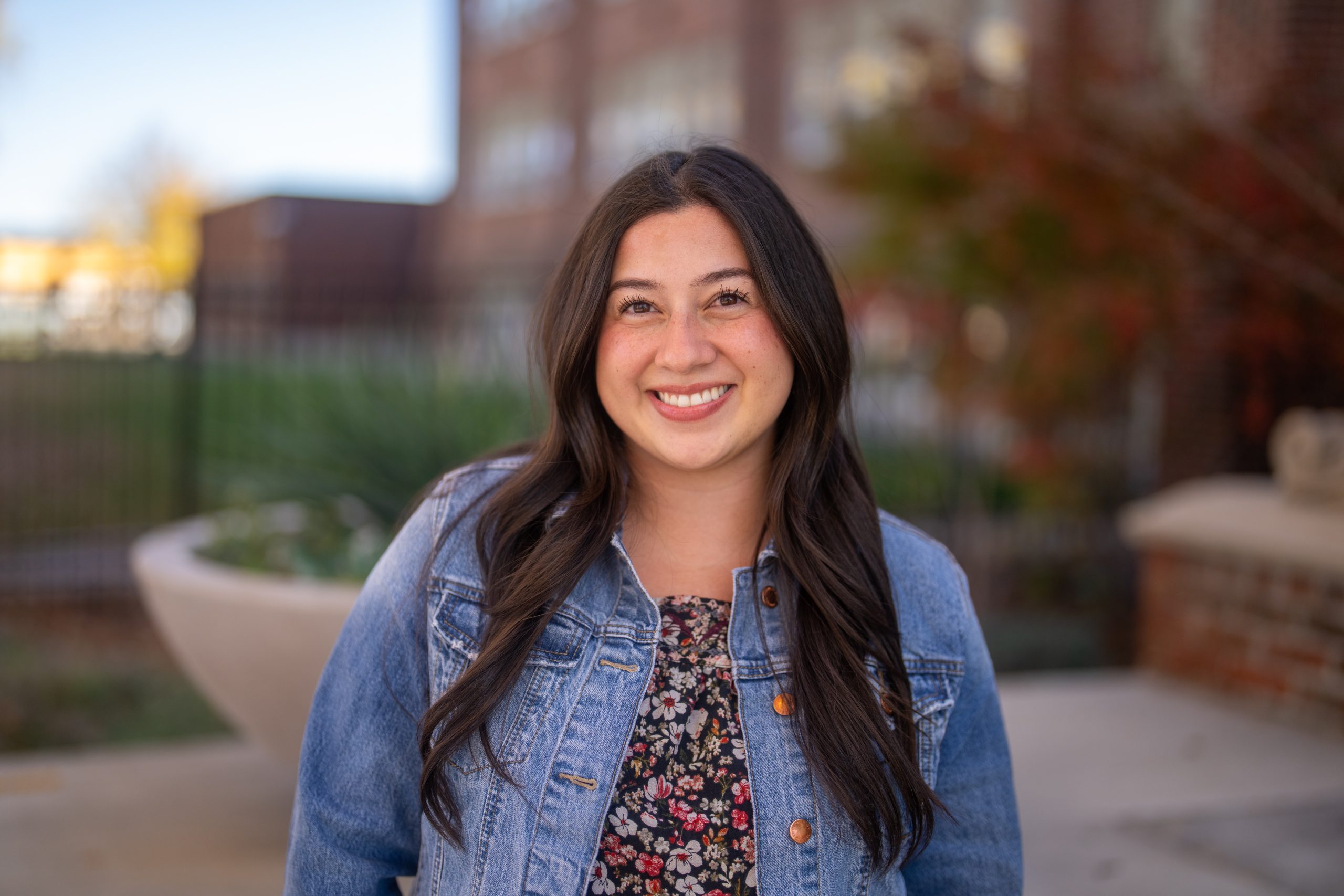 A smiling young woman with long dark hair wears a denim jacket over a floral dress, standing outdoors.