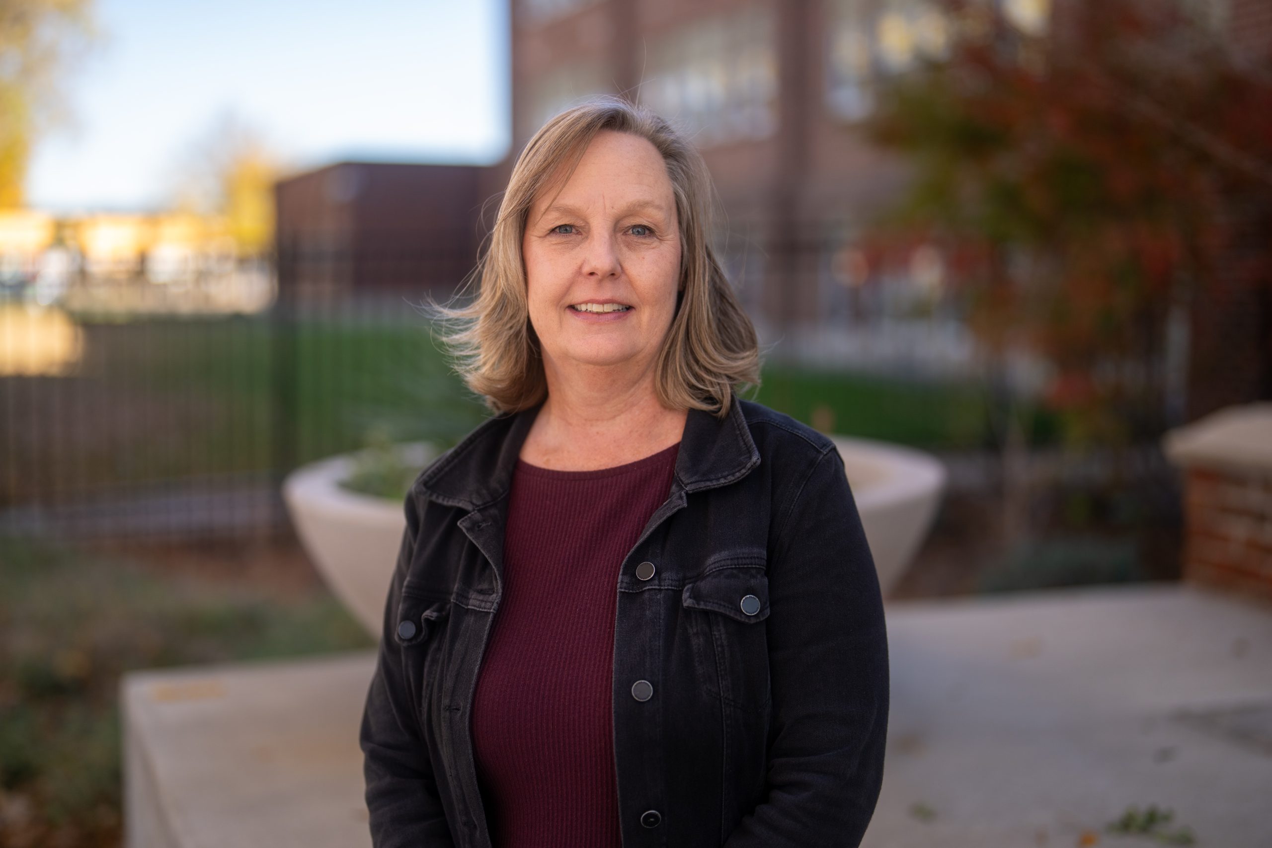 A woman with shoulder-length hair smiles while standing outdoors, wearing a black jacket and burgundy top.