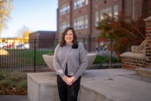 A woman stands on a ledge in front of a brick building, smiling, wearing a gray cardigan and black pants.