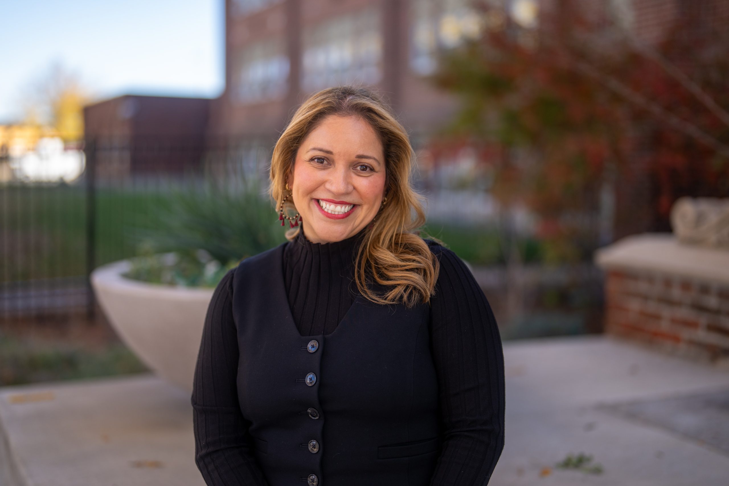 Smiling woman with long, wavy hair, wearing a black turtleneck and vest, outdoors with greenery in the background.