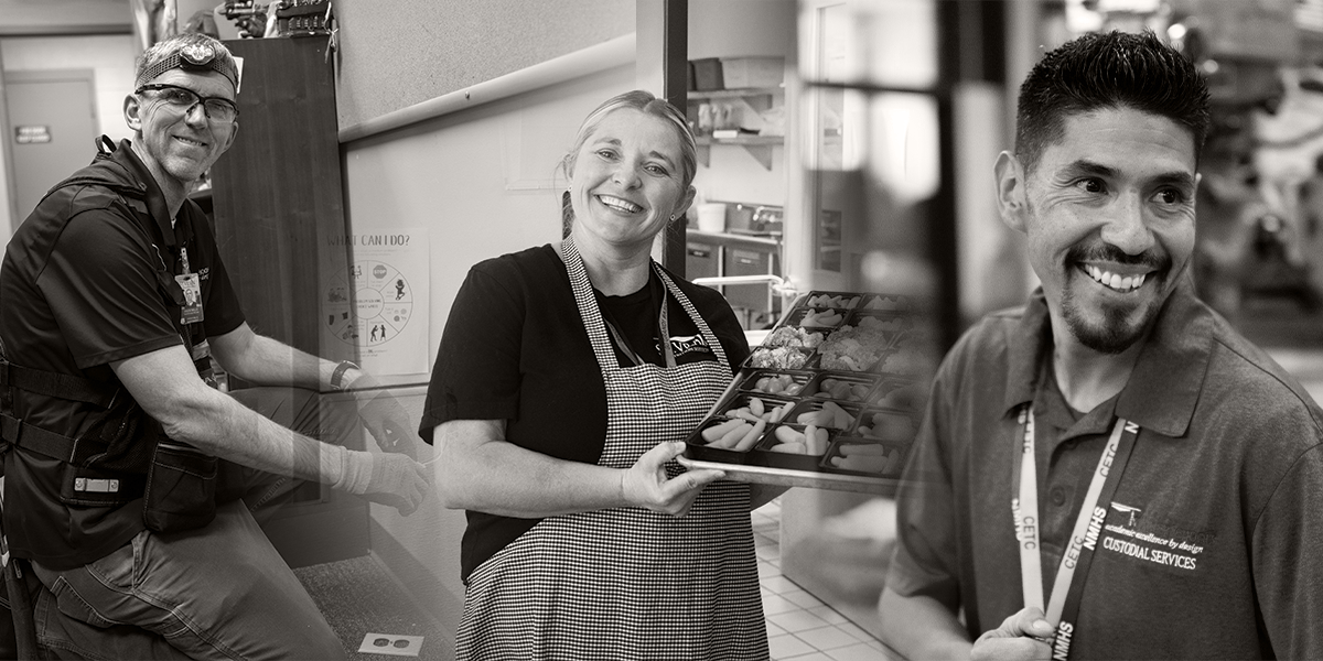 Three smiling individuals in a kitchen setting: one with a headlamp, one holding food trays, and another in a custodial uniform.