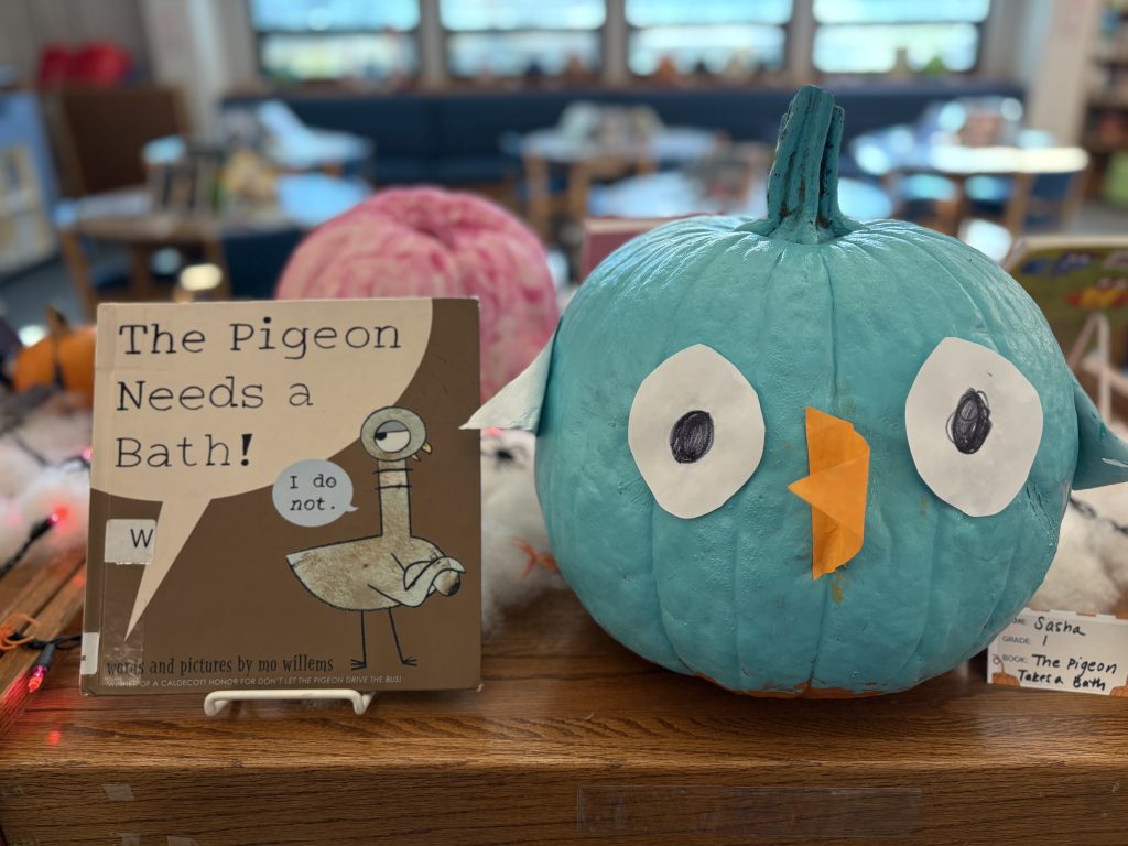 A teal pumpkin shaped like a bird sits beside the book "The Pigeon Needs a Bath!" on a wooden table.
