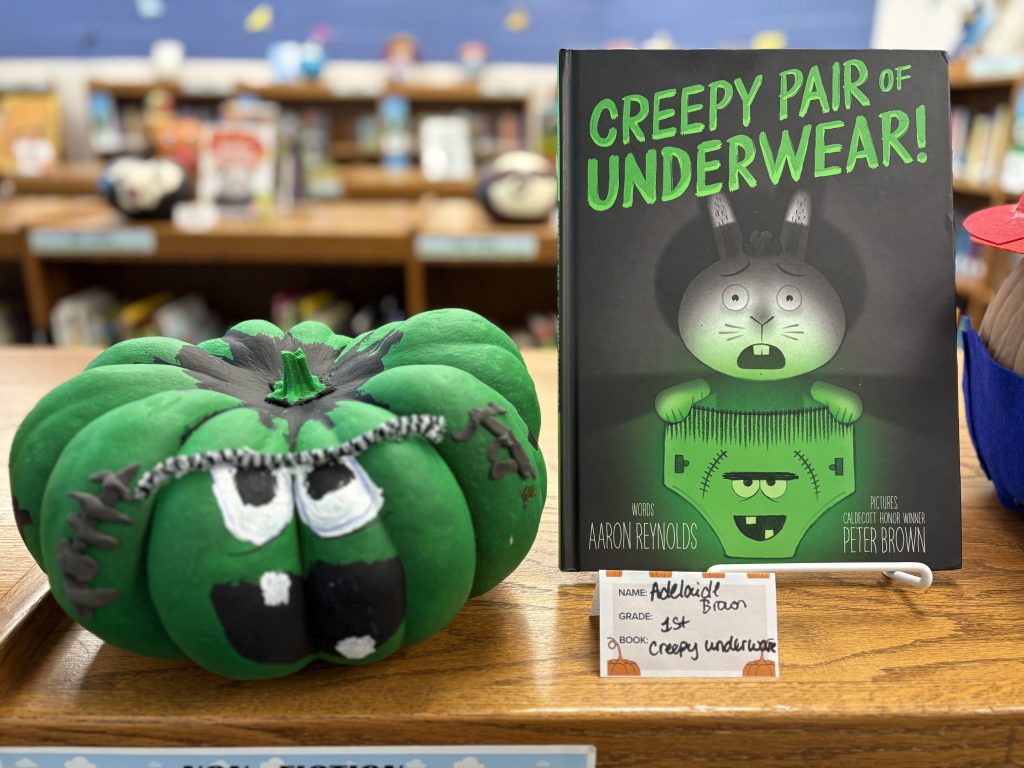 A green pumpkin with a face next to the book "Creepy Pair of Underwear!" by Aaron Reynolds on a library table.