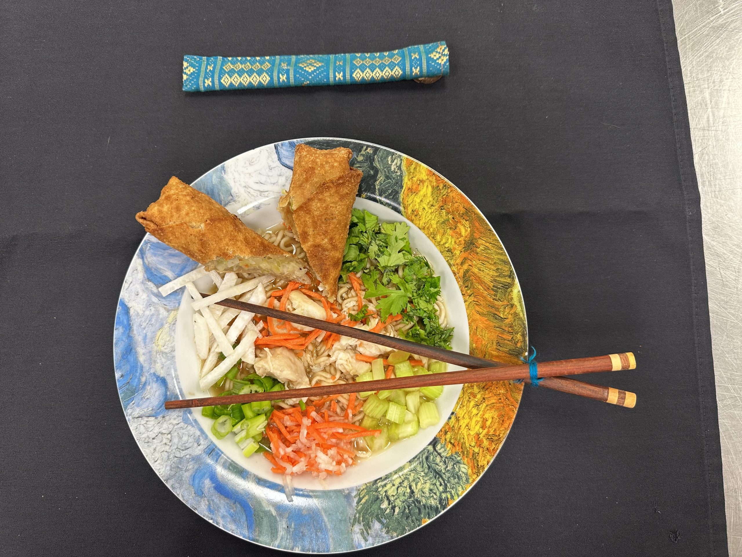 A colorful plate with noodles, vegetables, and two spring rolls, accompanied by chopsticks on a black tablecloth.