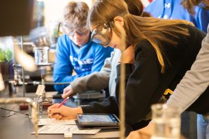 Students in safety goggles conduct an experiment, writing notes in a science classroom.