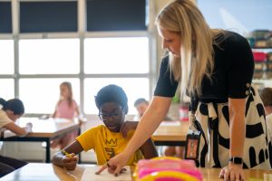 A teacher assists a student at a desk in a classroom filled with children engaged in various activities.
