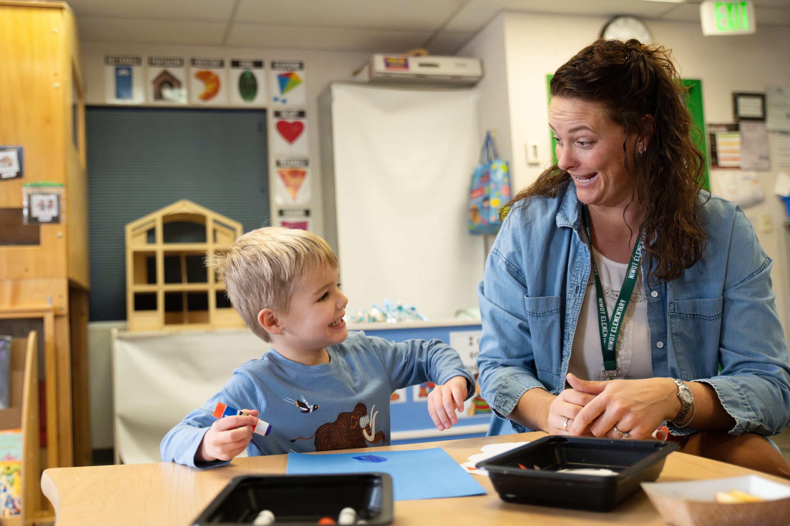 A smiling child and a teacher engage in arts and crafts at a classroom table, surrounded by educational materials.