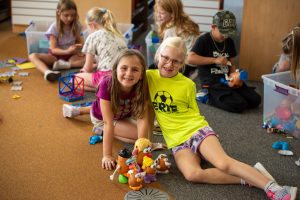 Two girls smile while sitting with a group of children playing with toys, including Mr. Potato Head, in a classroom.