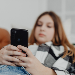 A young person lies on a couch, focused on their smartphone while wearing a checkered shirt.