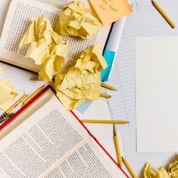 A cluttered workspace with open books, crumpled yellow paper, pencils, and a blank sheet on a table.