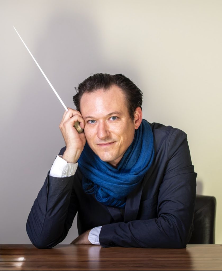 A man in a dark suit and blue scarf rests his chin on his hand, holding a conductor's baton.