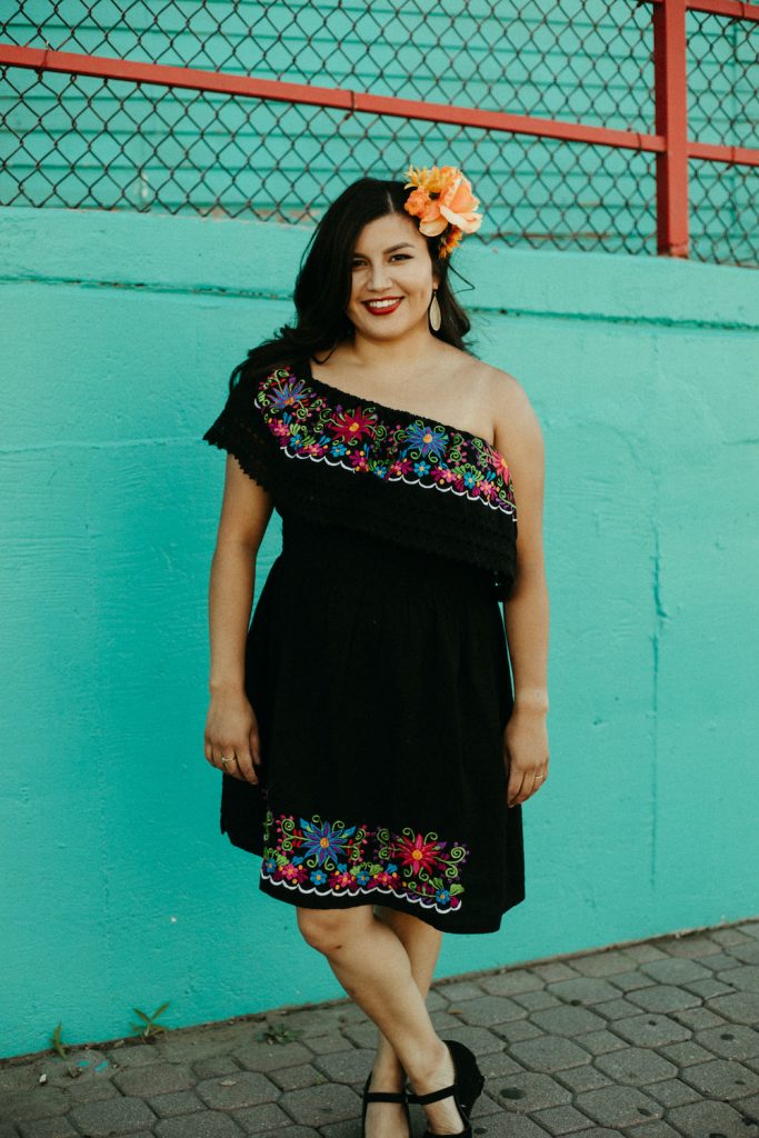 A woman in a black embroidered dress stands against a turquoise wall, smiling with a flower in her hair.