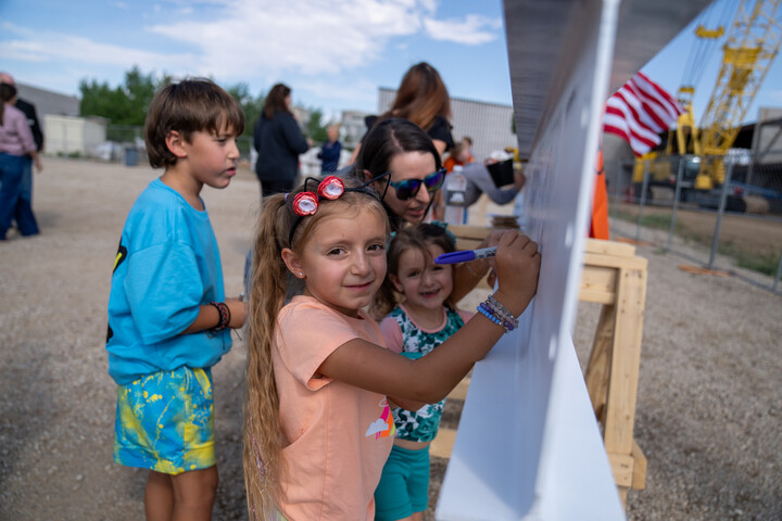 Children and adults participate in a community event, with kids writing on a large board outdoors.