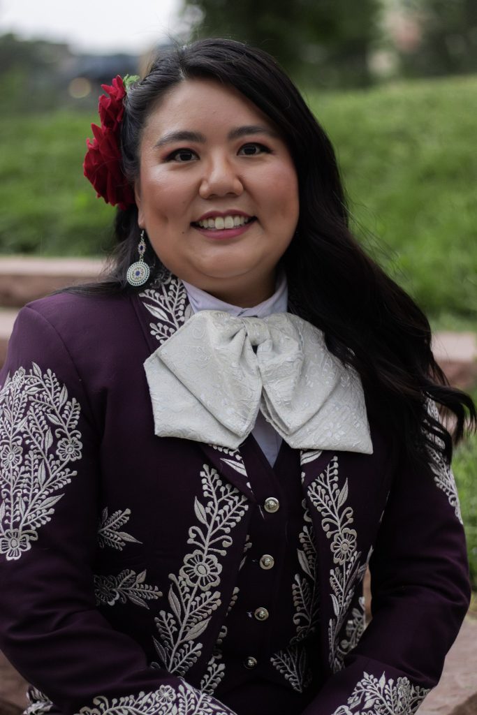 A woman in a purple mariachi suit with floral embroidery smiles, wearing a large bow and red flowers in her hair.