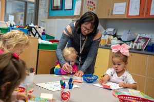 A teacher assists young children with coloring and drawing at a classroom table filled with art supplies.