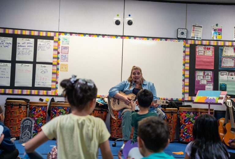 A music teacher plays guitar for a group of attentive children in a colorful classroom.