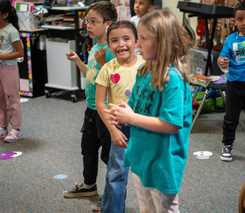 Three children are smiling and dancing in a classroom, engaging in a fun activity with colorful decorations around them.