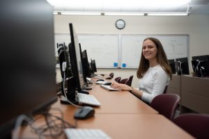 A student in a classroom smiles while sitting at a computer workstation, with multiple computers in the background.