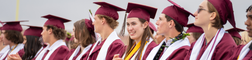 A row of graduates in maroon caps and gowns, smiling and celebrating during a graduation ceremony.