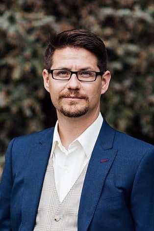 A man with glasses and a goatee wears a blue suit and white shirt, standing outdoors against a blurred background.