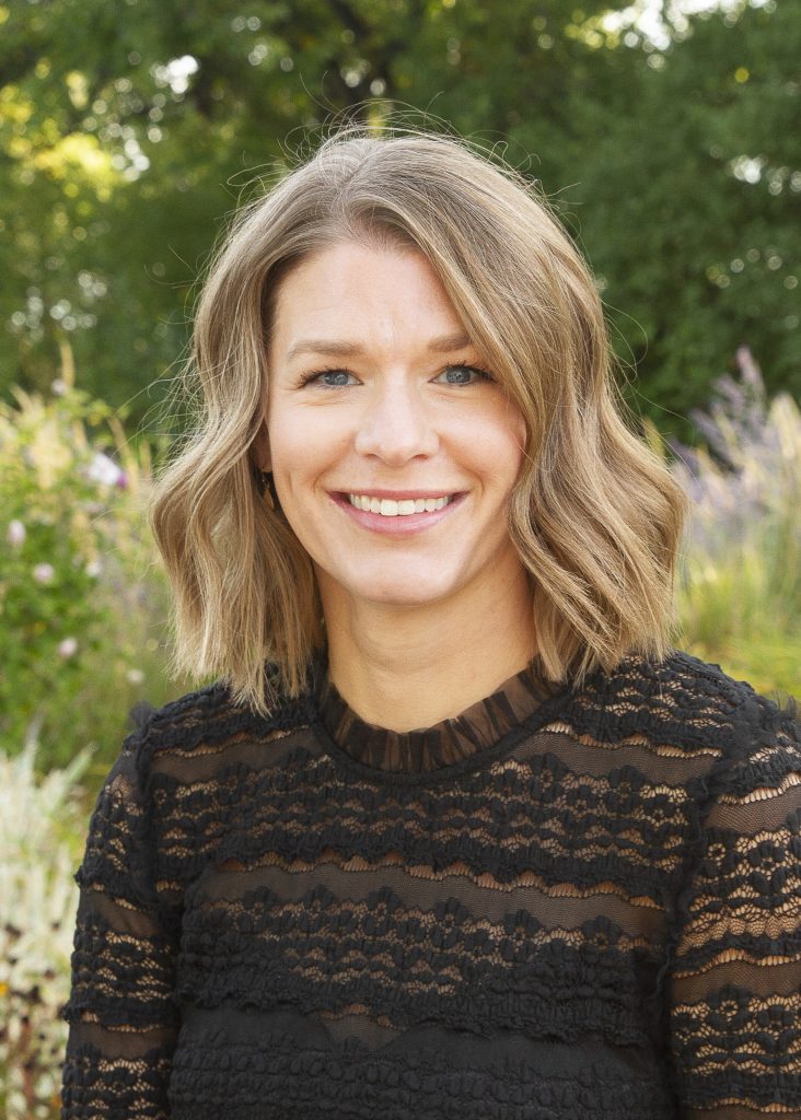 Smiling woman with wavy hair wearing a black lace top, standing outdoors with greenery in the background.