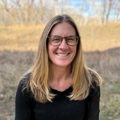A smiling woman with long blonde hair and glasses stands outdoors, with a blurred natural background.