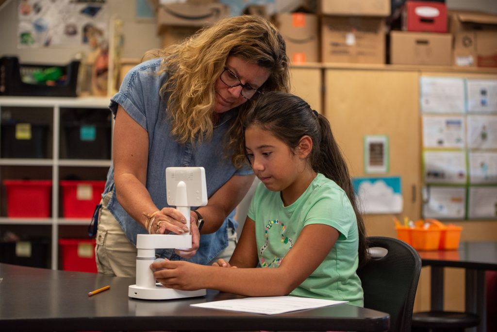 A teacher assists a young girl with a microscope at a classroom table, surrounded by educational materials.