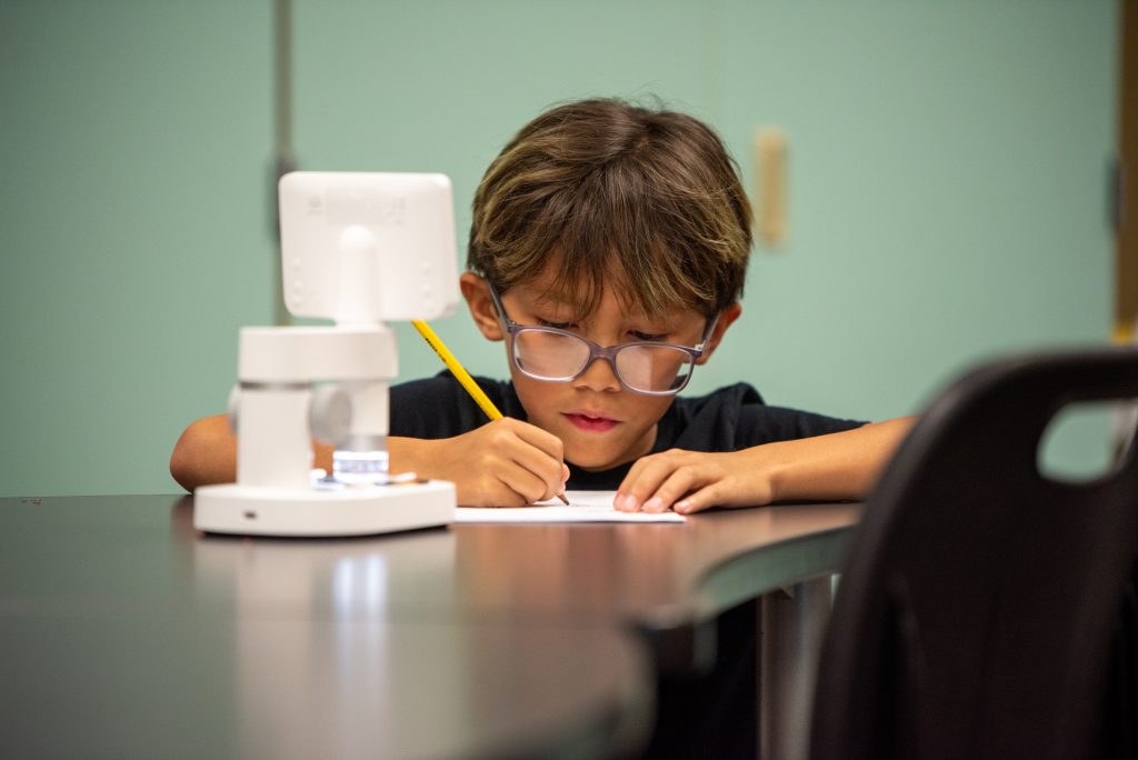 A young child with glasses writes with a pencil at a table, focused on their work, with a microscope nearby.