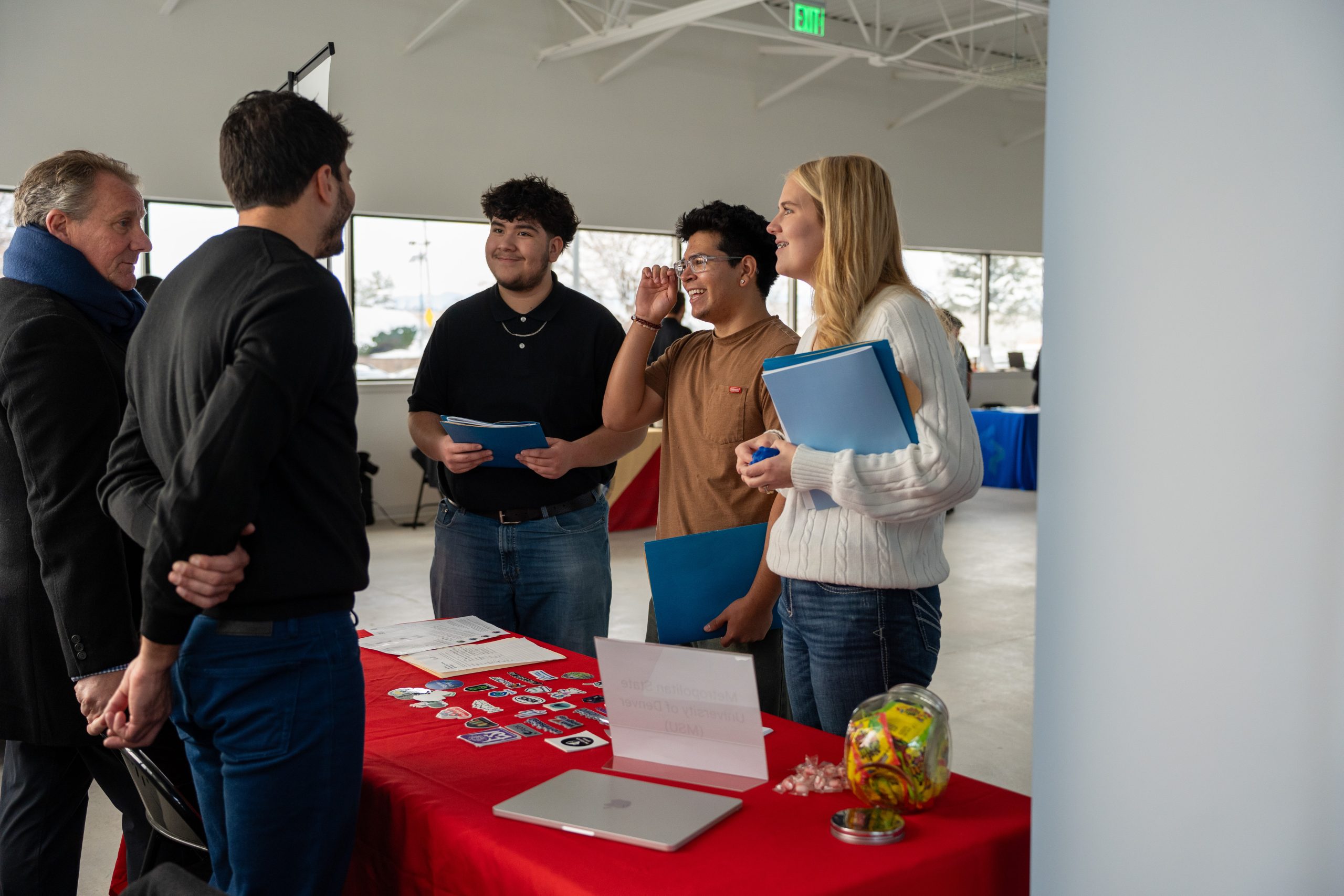 A group of four people engage at a table with a red cloth, displaying materials and snacks in a bright room.