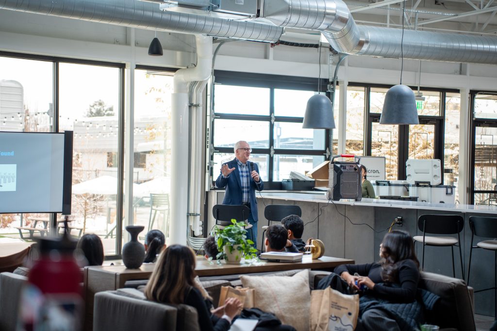 A speaker presents to an audience in a modern indoor space with large windows and casual seating.