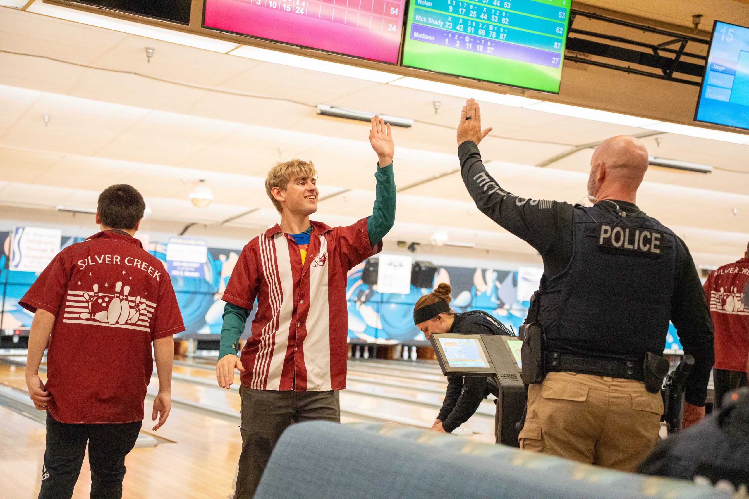 A teenager in a bowling alley high-fives a police officer, while others play in the background. Scores are displayed above.