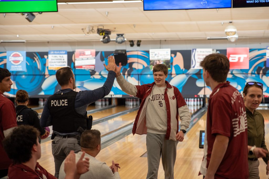 A police officer and a young man give each other a high five at a bowling alley, surrounded by cheering friends.