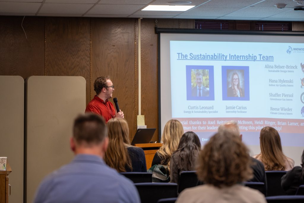 A speaker presents to an audience, discussing a sustainability internship team, with names displayed on a screen.