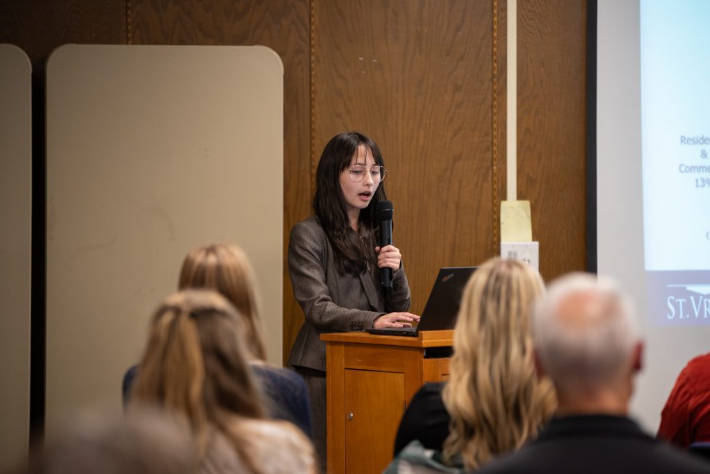 A student speaks at a podium with a laptop, presenting to an audience in a conference setting.