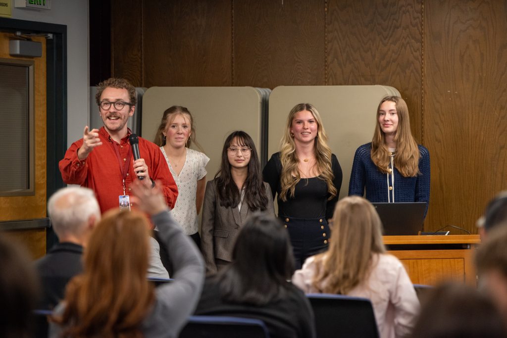 A group of five presenters stands on stage, engaging with an audience in a classroom setting.