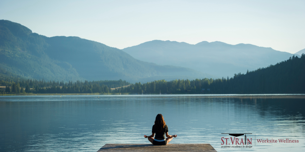 A person meditates on a dock by a calm lake, surrounded by mountains and trees under a clear sky.