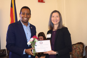Two people smile while holding a certificate and flowers at an event, with a decorative backdrop.