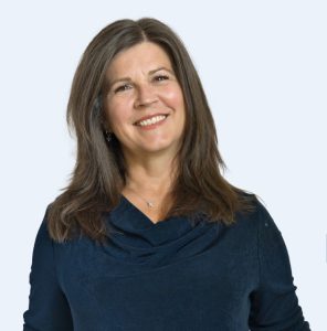 Smiling woman with long brown hair, wearing a dark blue top, set against a light blue background.