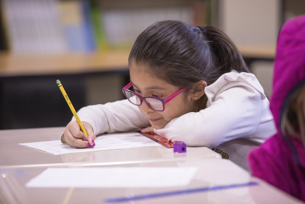 A young girl with glasses rests her chin on her hand while writing with a pencil on a worksheet at a desk.