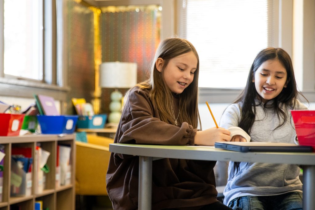 Two smiling girls are seated at a table in a classroom, engaged in writing and sharing ideas. Colorful bins are nearby.